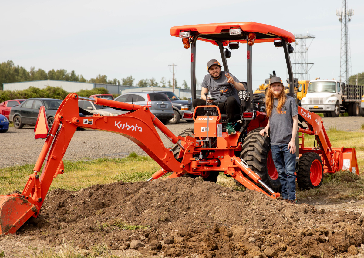 Two students posting with an orange Kubota backhoe. There is freshly dug ground near the bucket of the backhoe, indicating the students have been operating it.